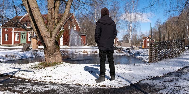 Gabriel står med ryggen mot kameran i ett snöigt landskap. I bakgrunden syns flera röda hus, en gärdesgård och en liten damm.