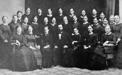 Svartvitt arkivfoto med cirka 30 kvinnor klädda i mörka klänningar, i mitten av gruppen sitter en ensam man.