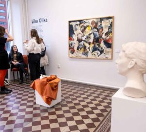 Vid en utställning syns en grupp personer samtala i ett hörn. På väggen hänger en stor tavla. På det schakrutiga golvet står två piedestaler. Den mellangrunden en med två keramikhänder som vilar på ett orange tyg. På piedestalen i förgrunden står en keramikbyst.