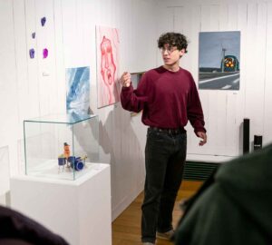 En man står framför flera tavlor och en glasmonter med ett keramikverk. Han visar och berättar om dem för publiken.
