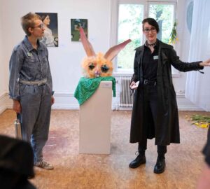 Två personer står på var sin sida av ett stort kaninhuvud på en piedestal. De berättar om utställningen för flera åhörare.