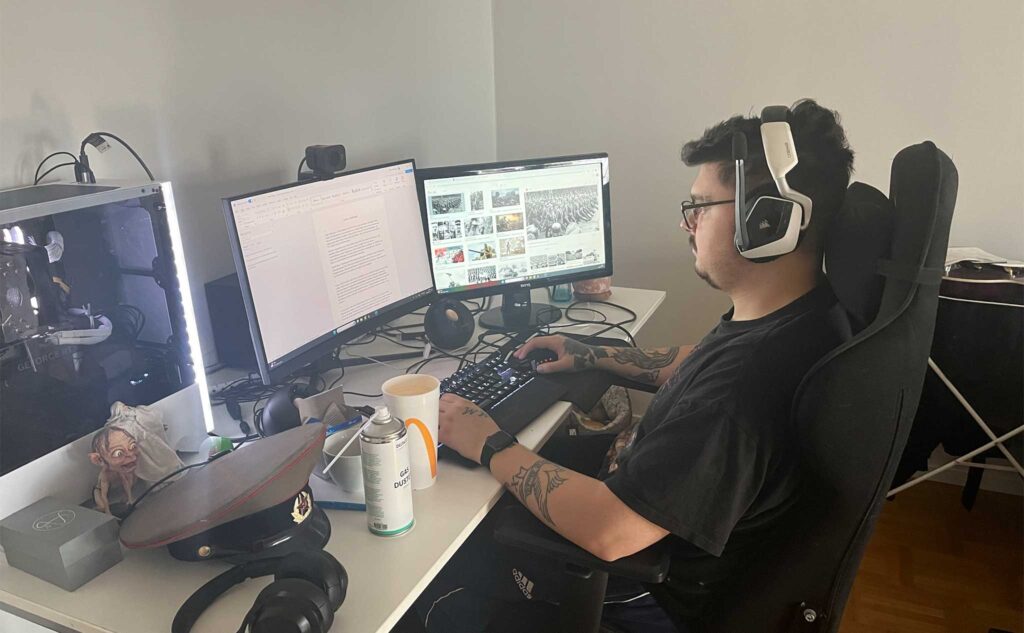 Ung man med svart t-shirt, mörkt hår och glasögon i sidoprofil. Sitter vid skrivbord med dator och dubbla bildskärmar.