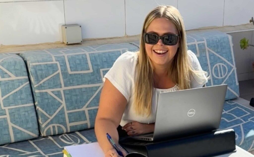 Ung kvinna med långt ljust hår, solglasögon och vit t-shirt sitter utomhus vid bord med laptop och anteckningsmaterial.